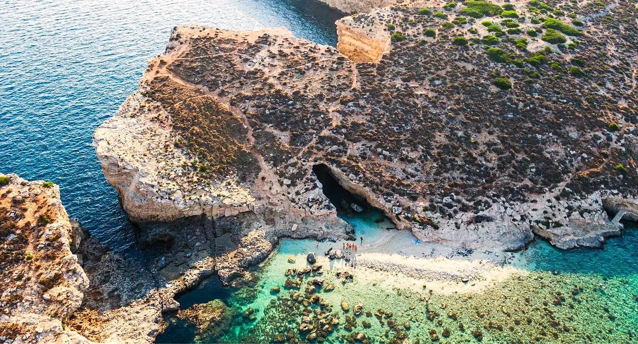 La pequeña cala de arena de Cominotto desde el aire, con una cueva en la roca y el agua turquesa