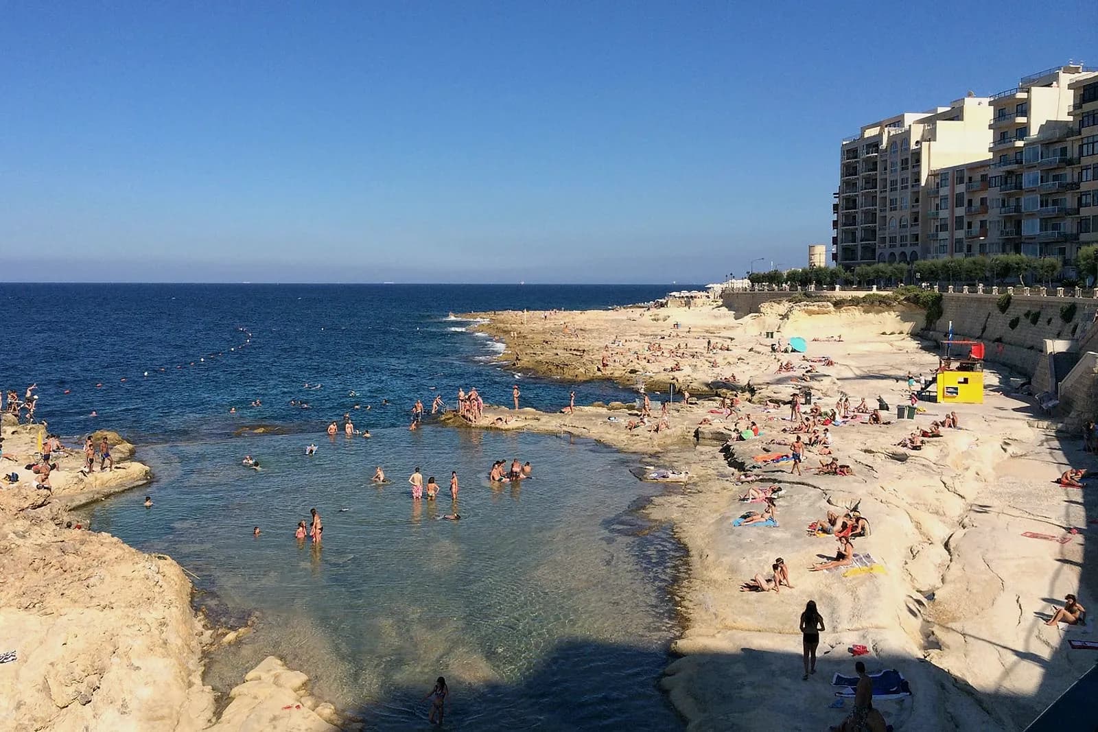 La bahía de Fond Għadir, en Sliema, con bañistas en la cala de agua clara, la roca llena de toallas y la caseta de socorristas