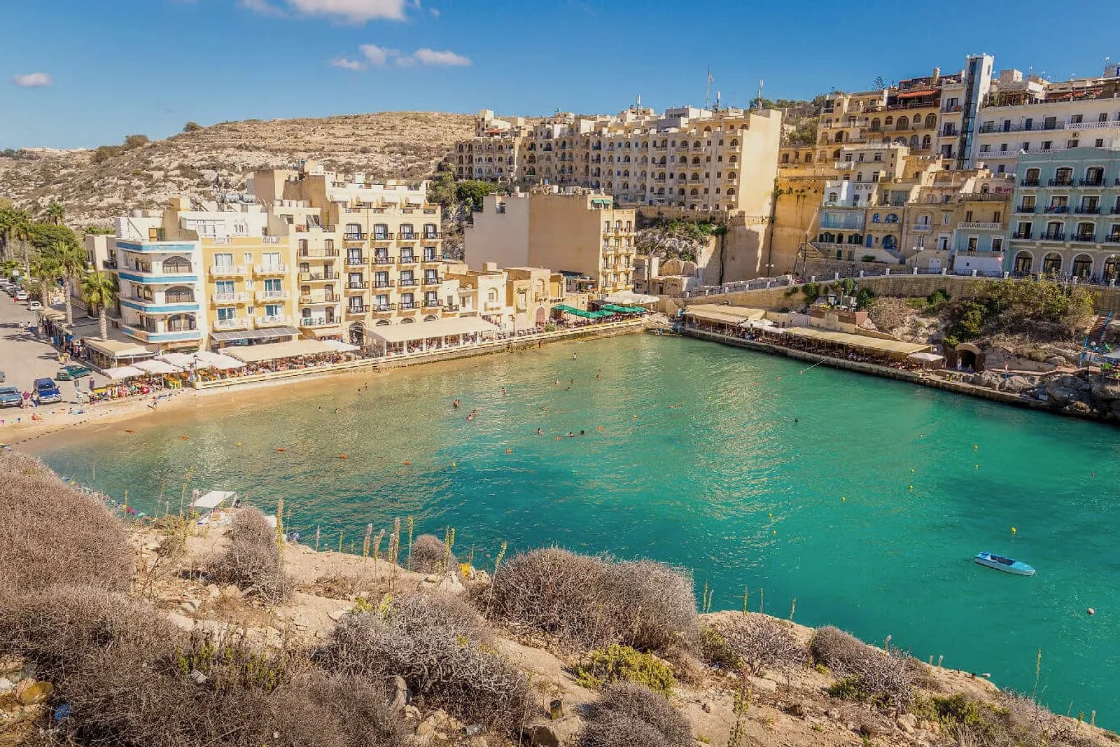 La bahía de Xlendi, con el agua turquesa, la playa al fondo y los edificios del paseo